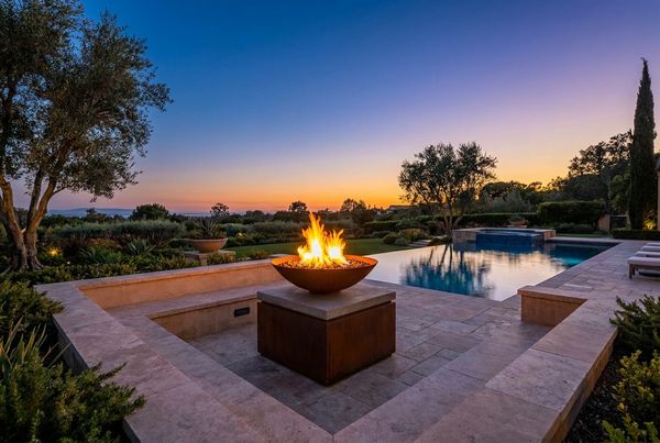 Fire bowl on a slender pedestal glowing at dusk beside a modern pool